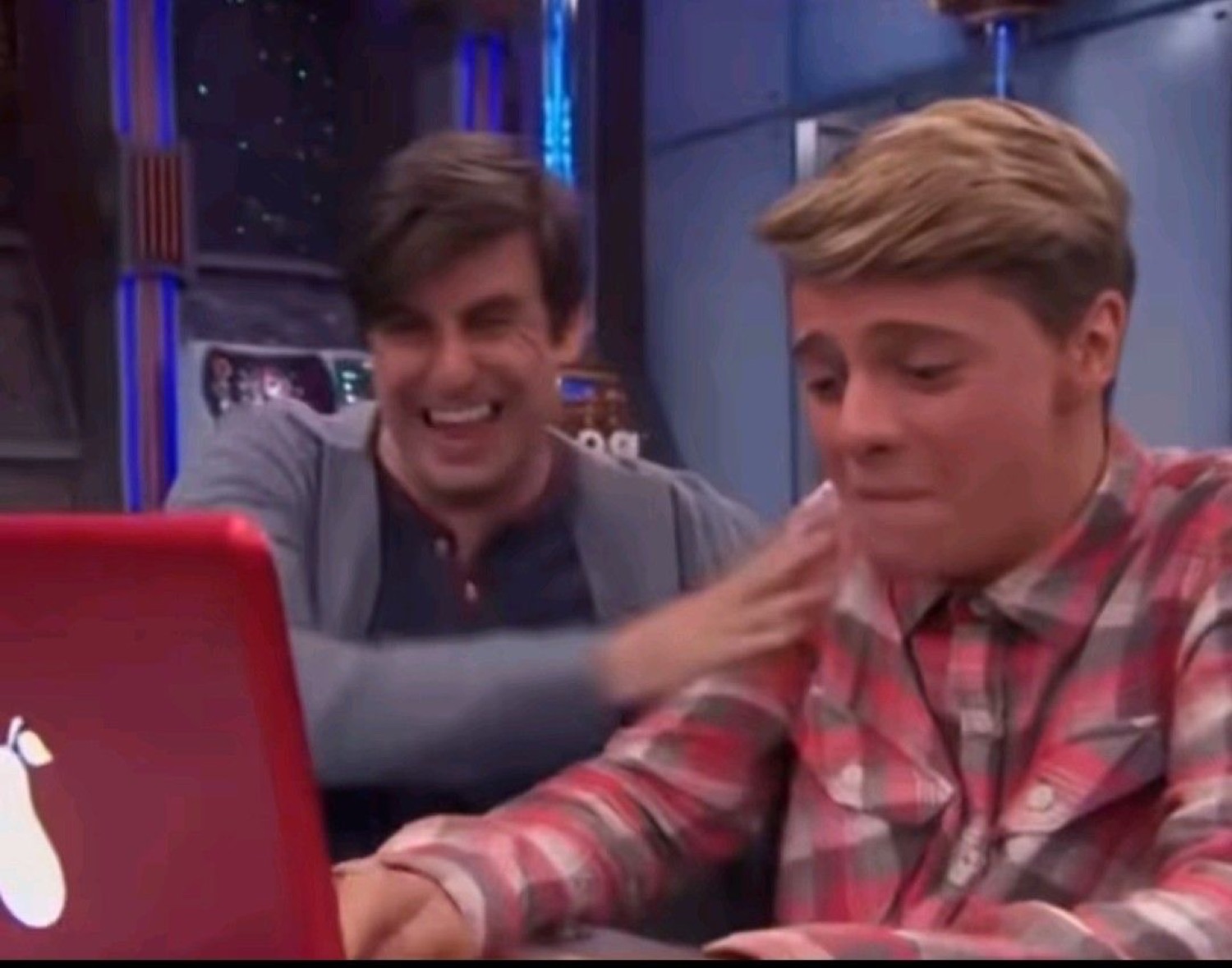 One guy laughing while patting a second guy's shoulder who looks uncomfortable at a laptop