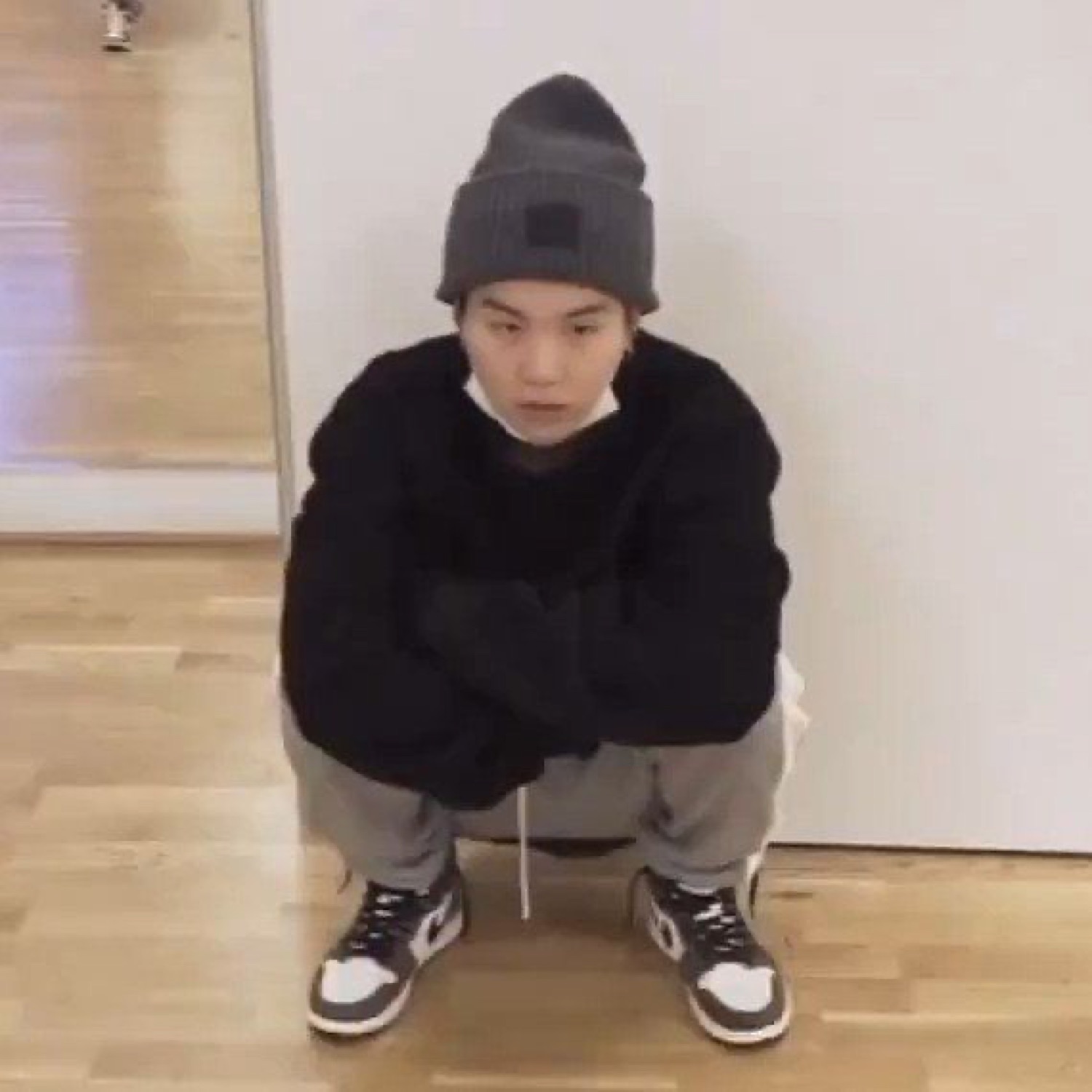 Guy in a beanie and black sweater squatting on the floor with a flat, blank stare