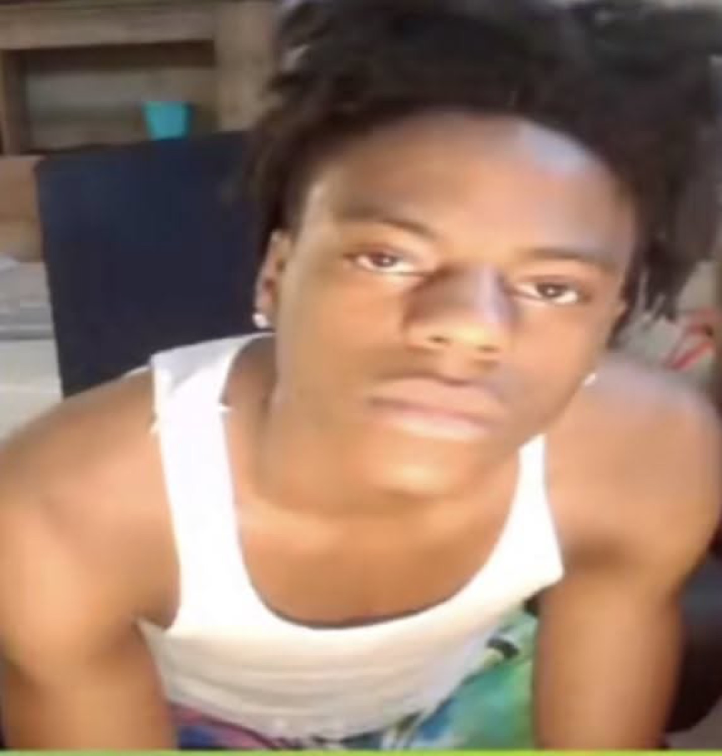 Young man in a white tank top leaning toward the camera with a flat, deadpan face