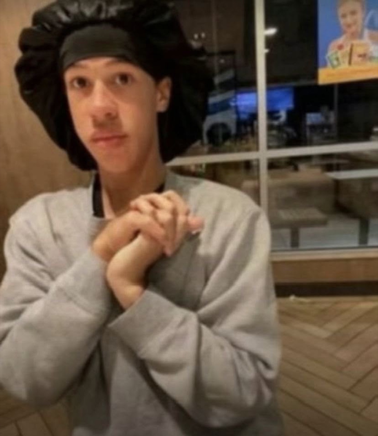 Young person in a satin bonnet standing in a fast-food restaurant with hands clasped nervously