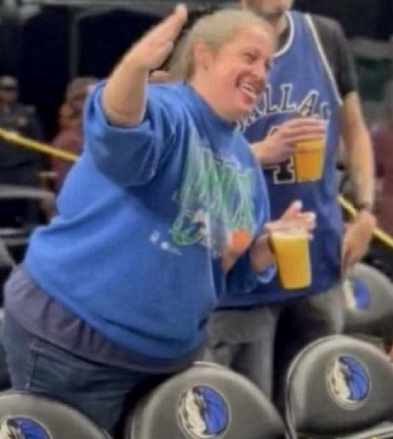 Woman in a Dallas Mavericks shirt dancing at a game with a drink in hand