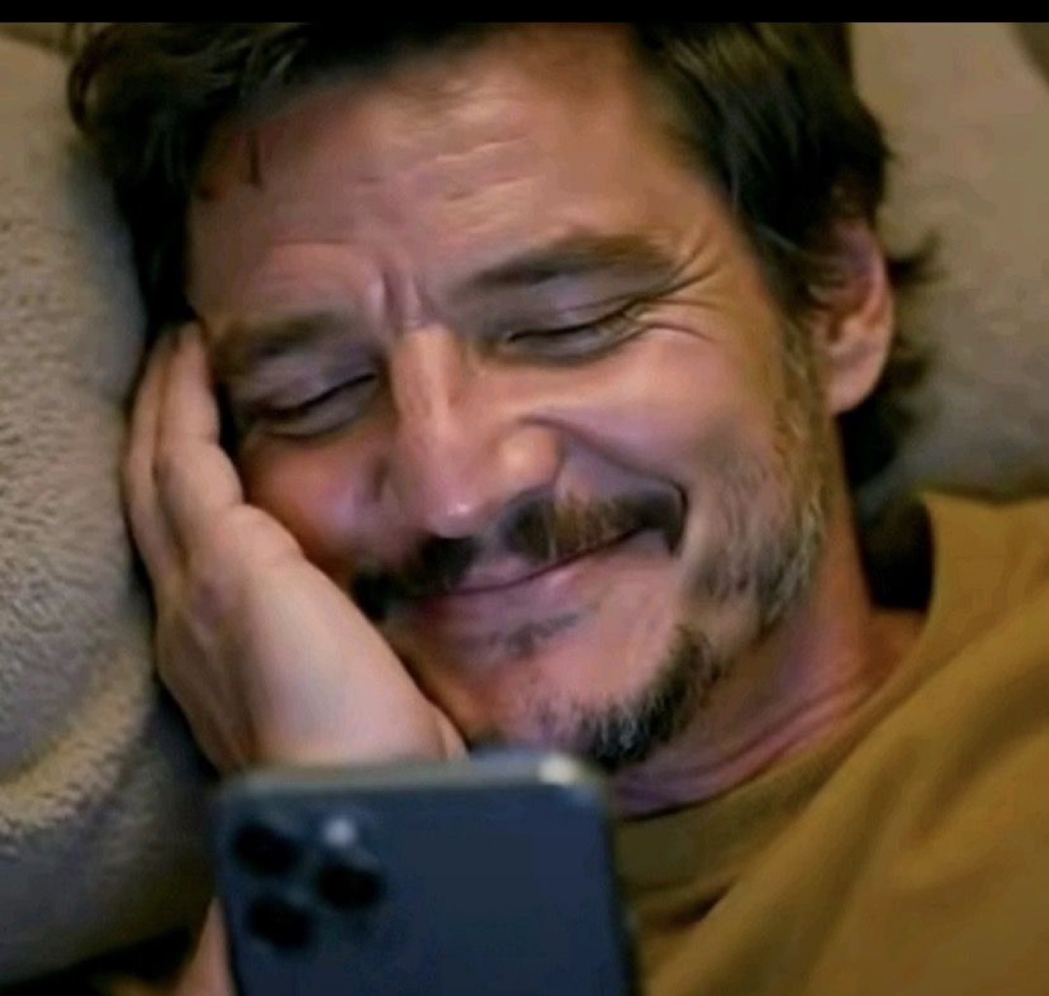 Pedro Pascal lying down, smiling softly at his phone with a cozy, smitten look