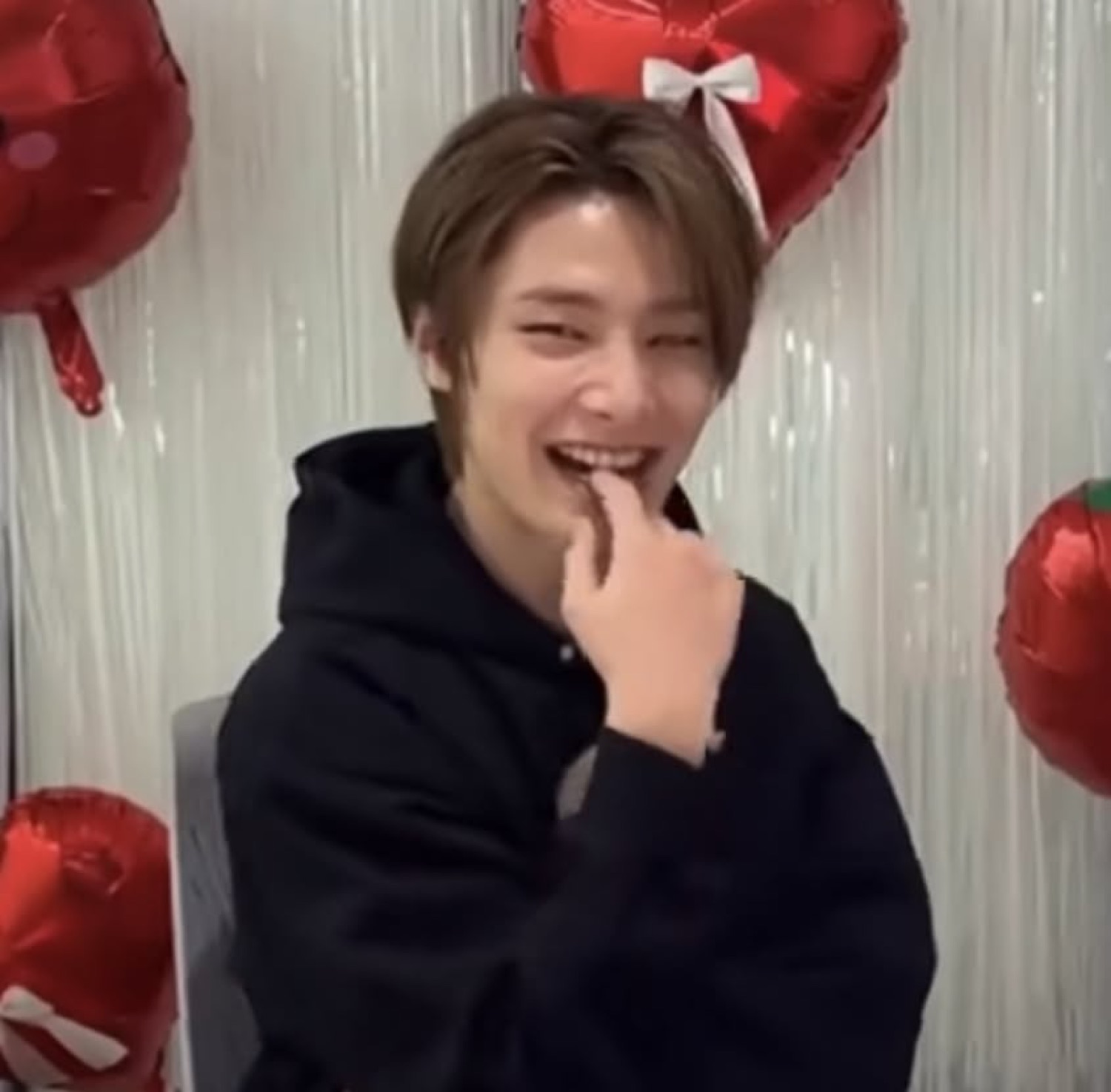 Guy in a black hoodie giggling into his hand in front of red heart balloons