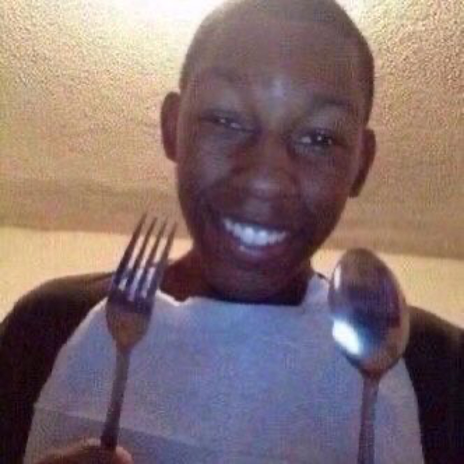 Kid in a napkin bib holding up a fork and spoon, grinning like dinner is served