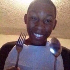 Kid in a napkin bib holding up a fork and spoon, grinning like dinner is served