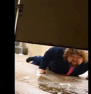 Curly-haired man sprawled face-down on wet pavement, still smiling at the camera