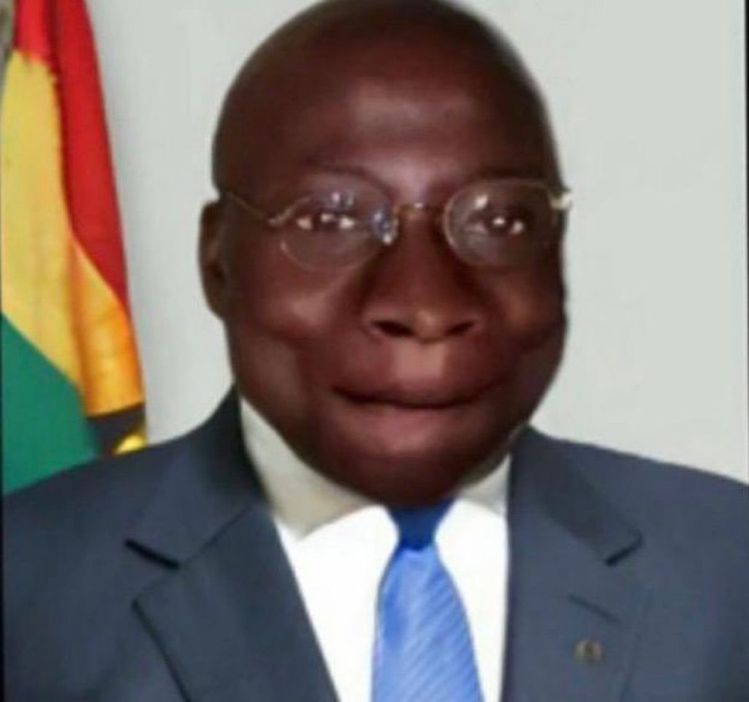 Man in a suit and glasses giving a stiff, tight-lipped half smile in front of a flag