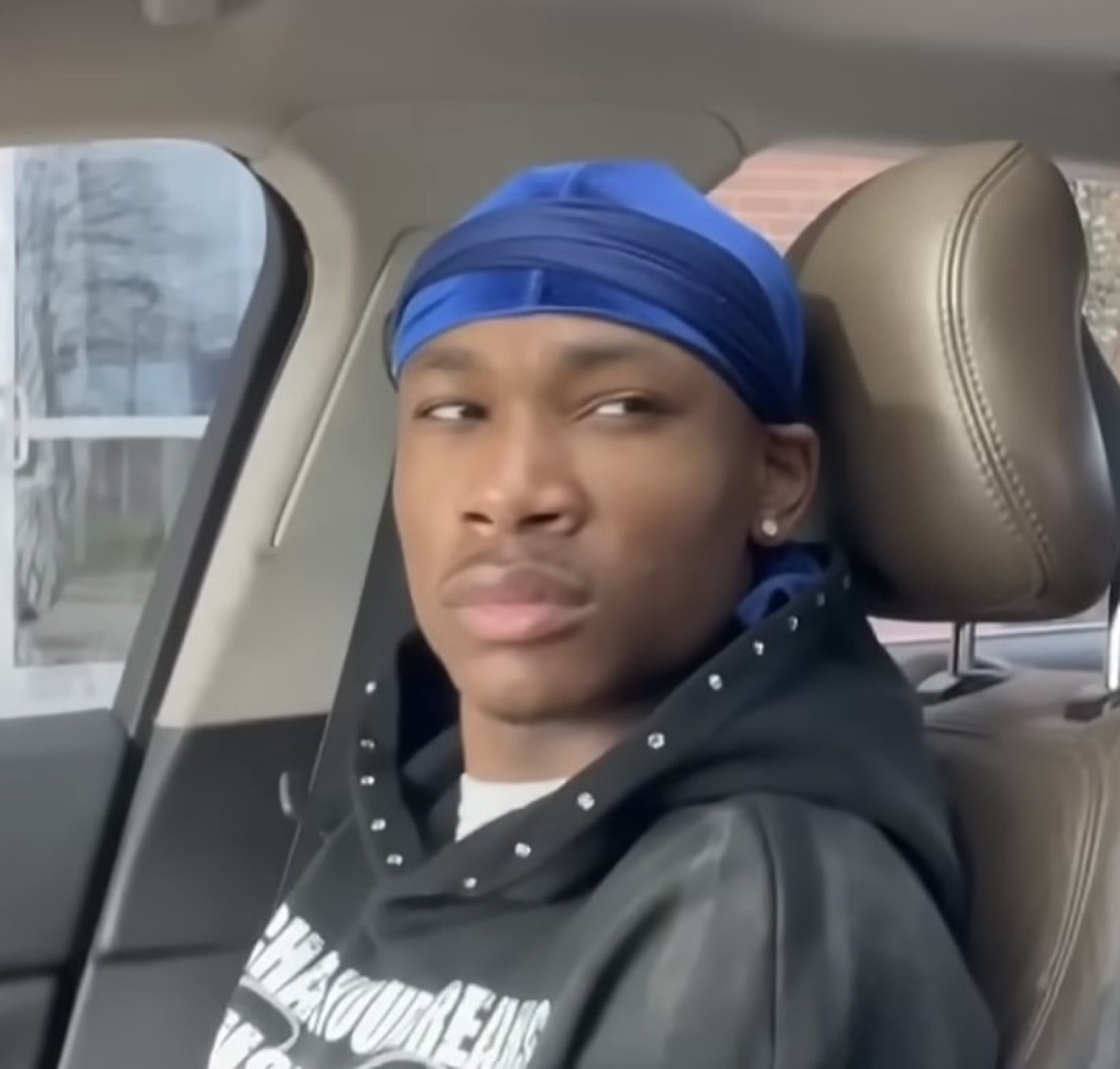 Young man in a blue durag leaning back in a car seat with a skeptical side-eye