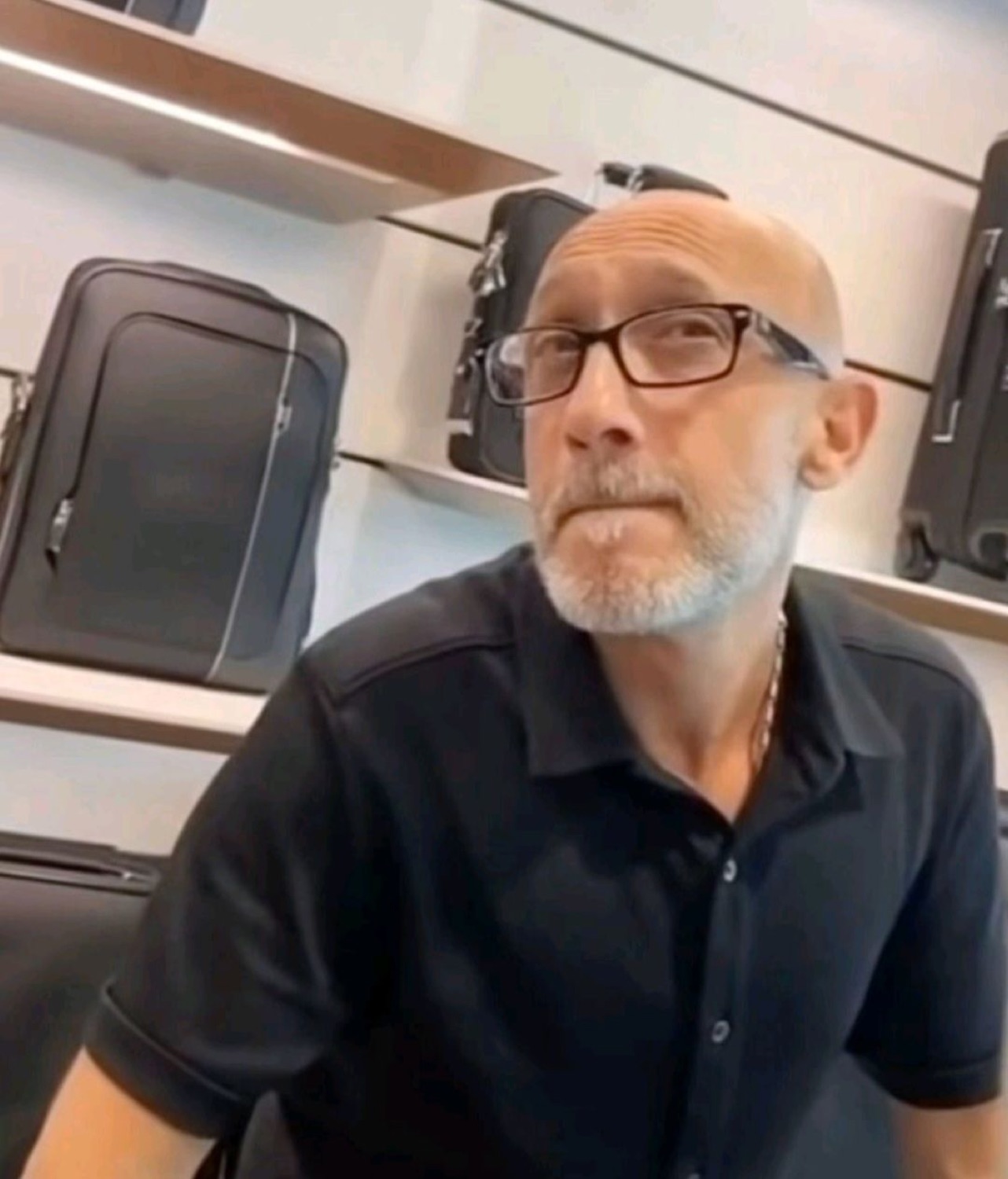 Bald man in glasses seated in a bag store staring flatly, thoroughly unimpressed