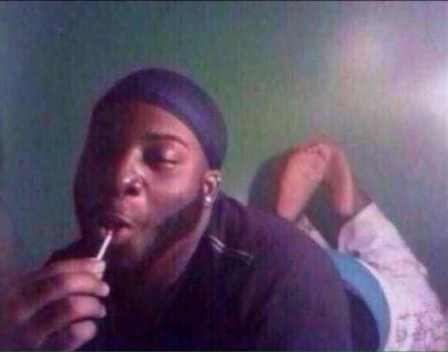 Man in a durag calmly lighting up while someone sprawls out behind him