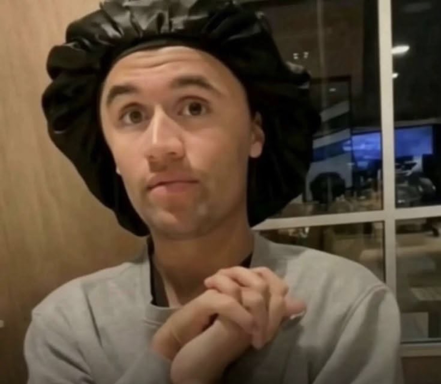 Guy in a silk bonnet clasping his hands with a coy, hopeful smirk