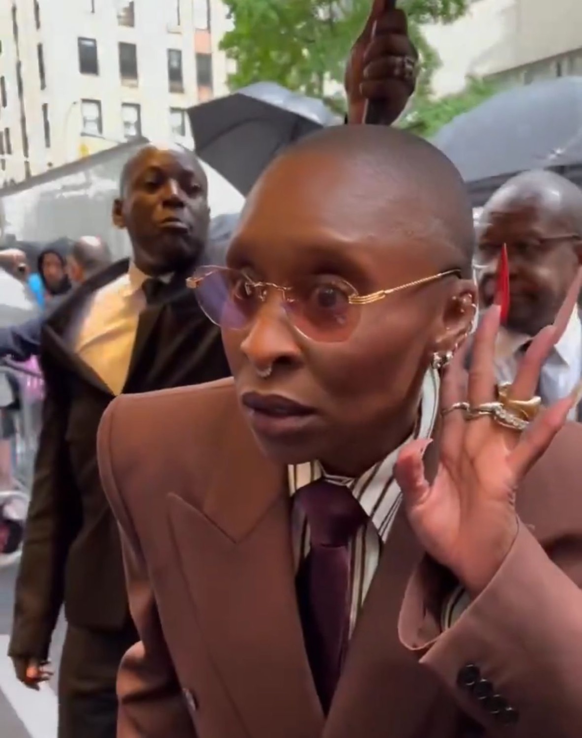 Bald woman in a brown suit raising one hand with a skeptical unimpressed sideways look