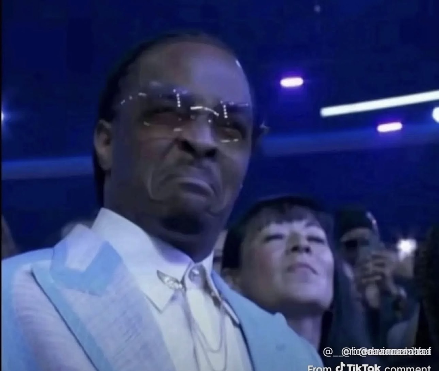 Man in a white suit and sunglasses at a blue-lit event, looking unimpressed