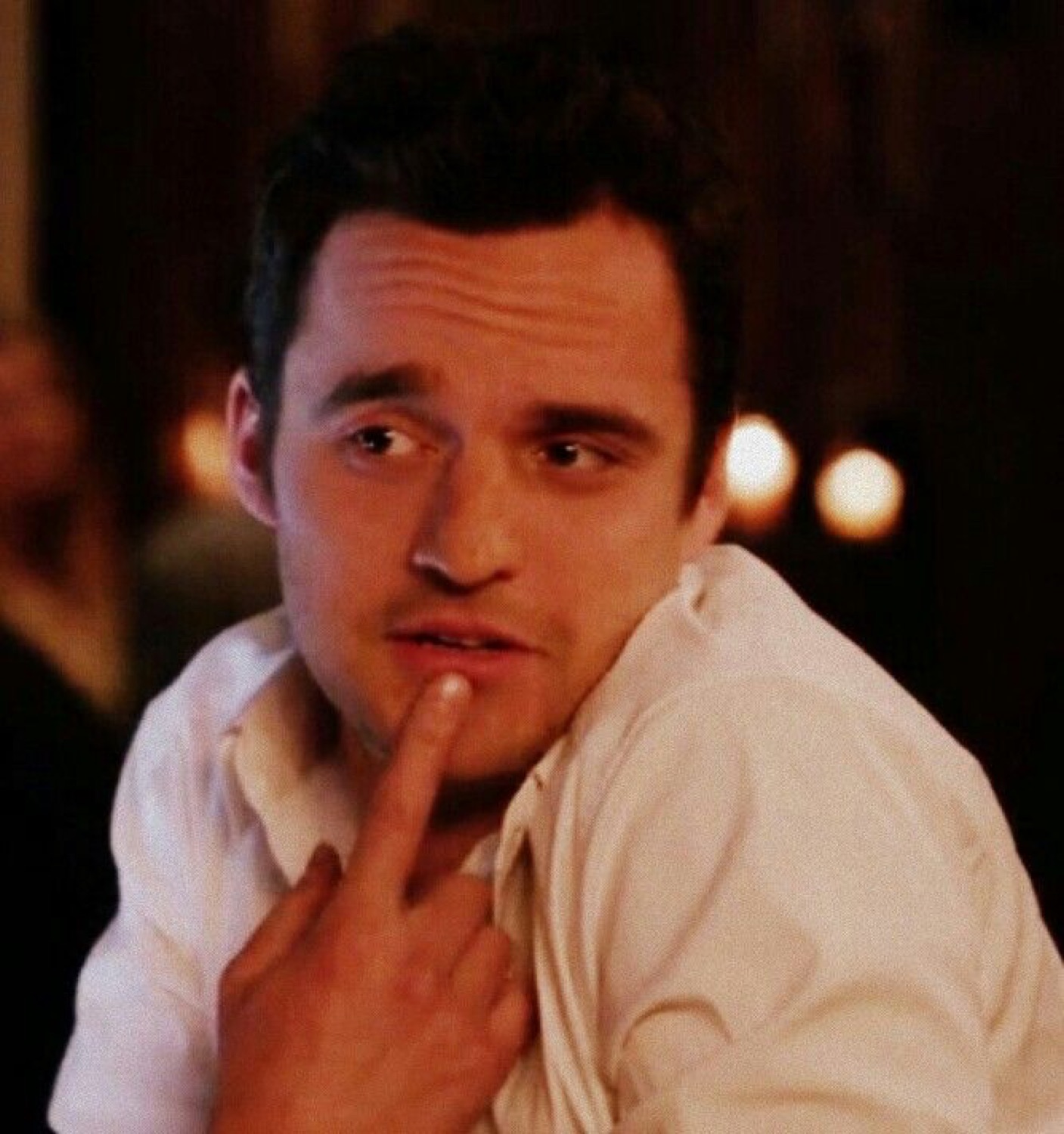 Man in a white shirt resting his finger on his lip with a calculating sideways glance