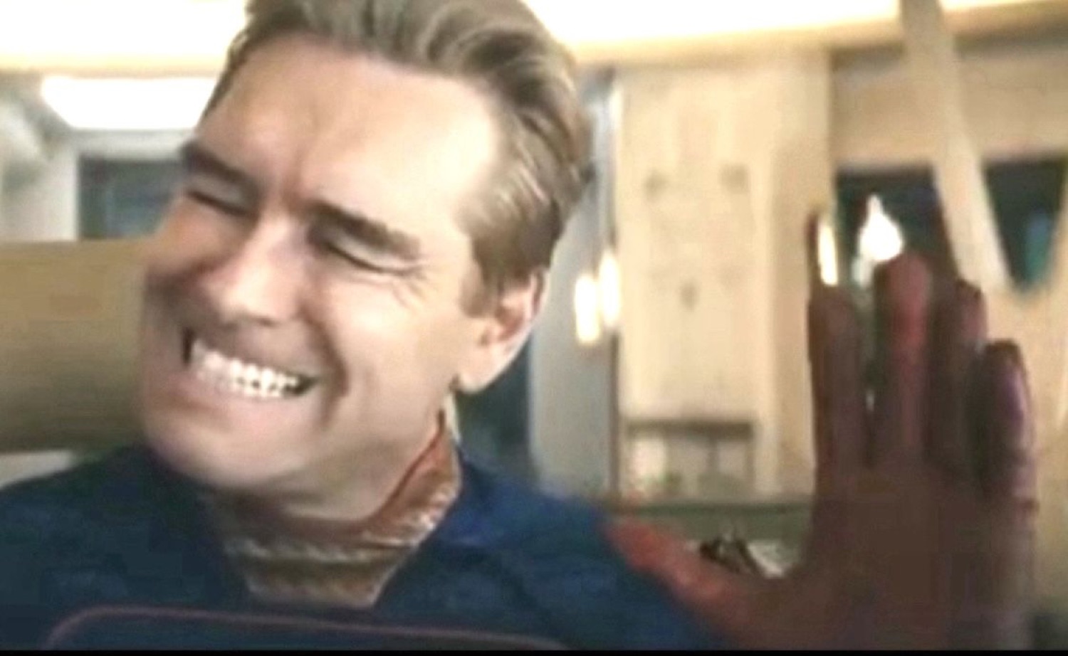 Blond man in a superhero suit forcing a pained, awkward laugh with clenched teeth