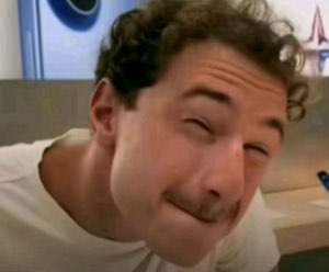 Curly-haired man leaning in close with squinting eyes and a smug, mocking little grin
