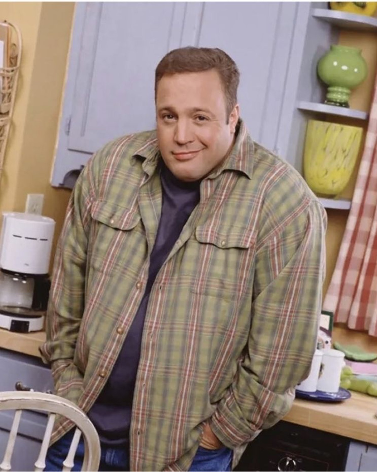 Man in a plaid shirt leaning against a kitchen counter with a small awkward smirk