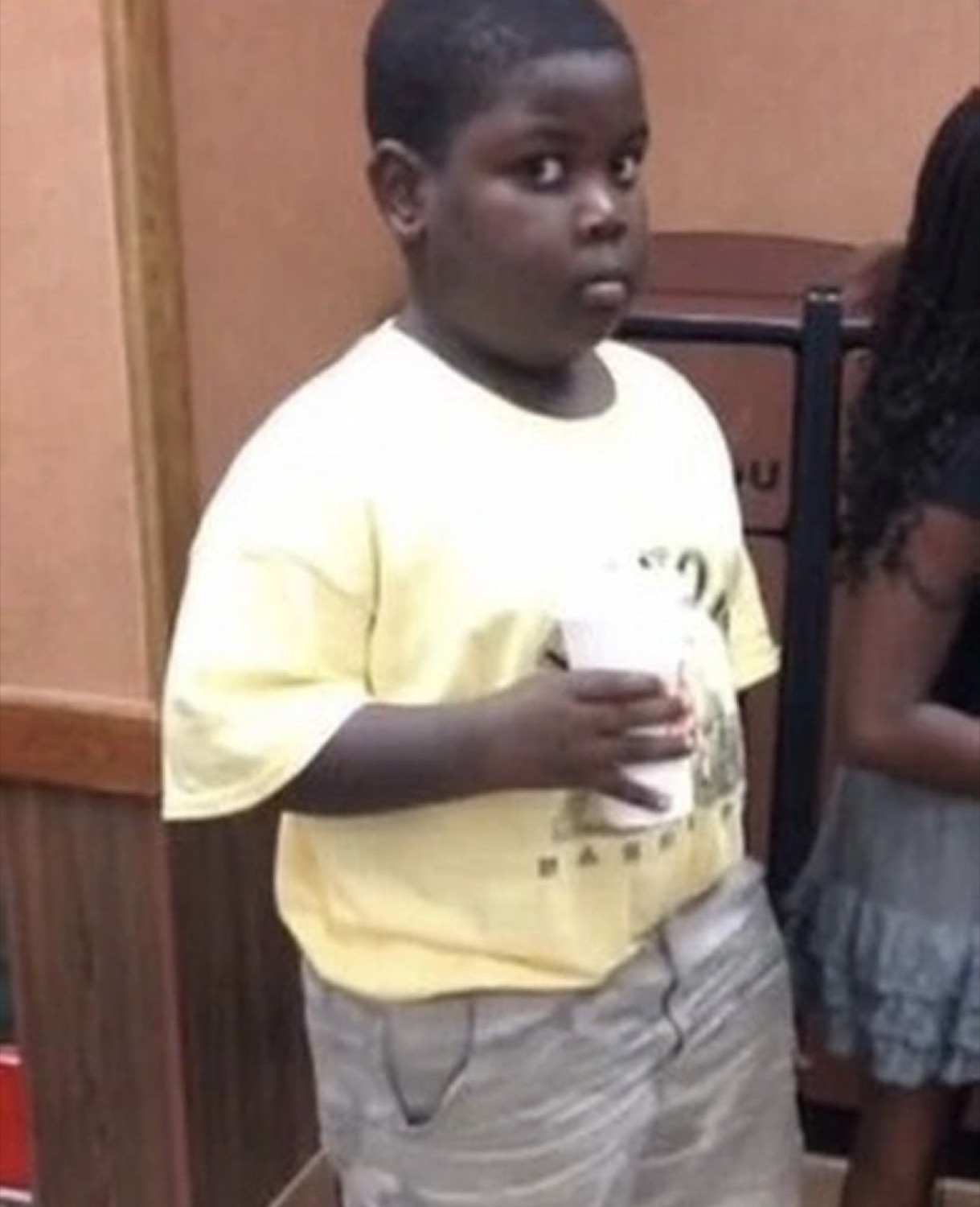 Kid in a yellow tee clutching a drink cup, glancing off with a caught-out look