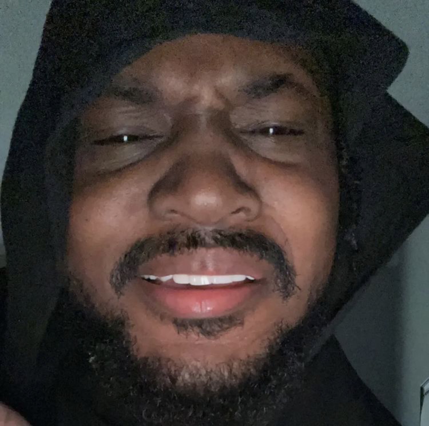 Man in a hood leaning into the camera with a squinting, smug grin