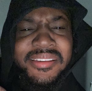 Man in a hood leaning into the camera with a squinting, smug grin