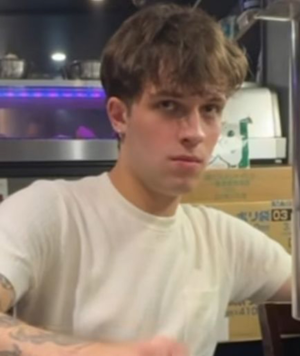 Tattooed guy in a white tee at a bar looking at something with total flat indifference