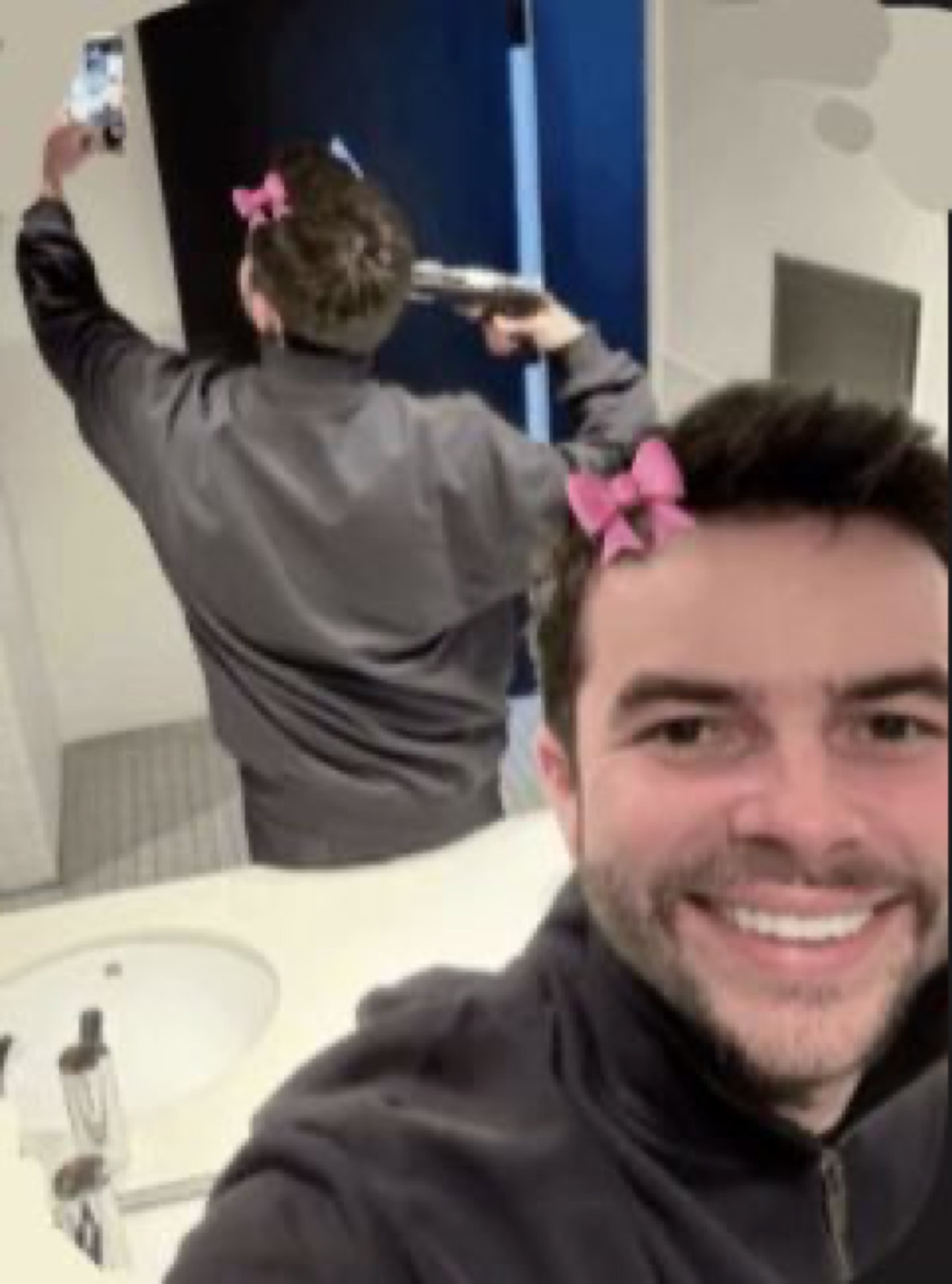 Grinning selfie with a friend in pink hair bows filming in a mirror behind him