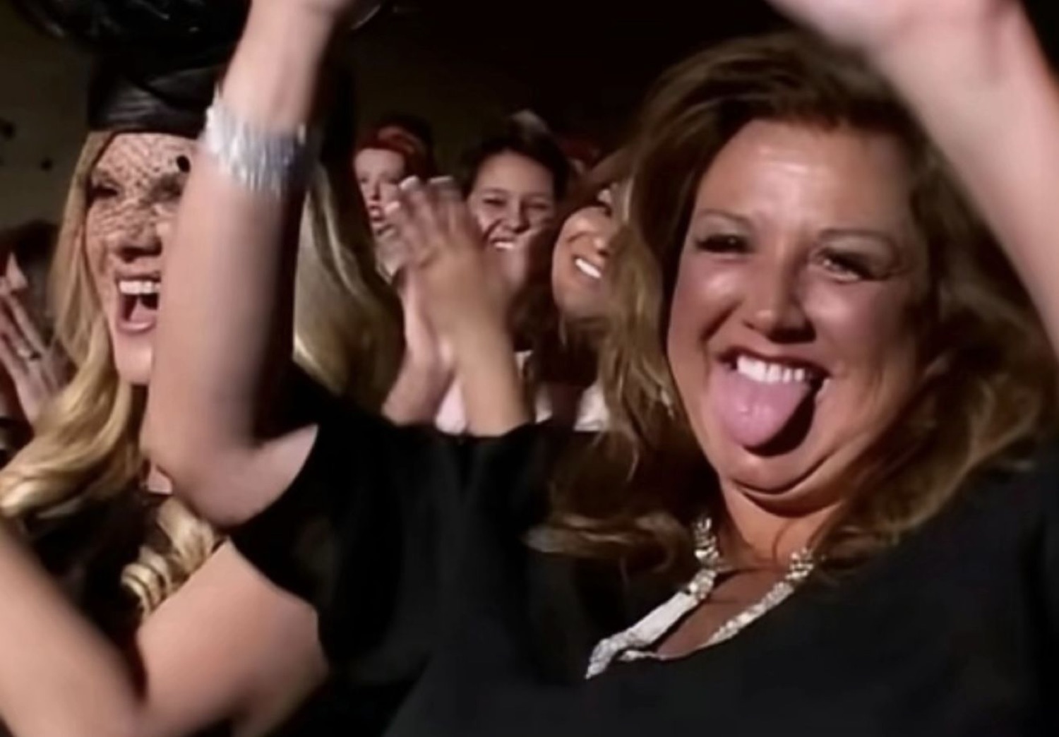 Woman in a crowd clapping and sticking her tongue out mid-celebration