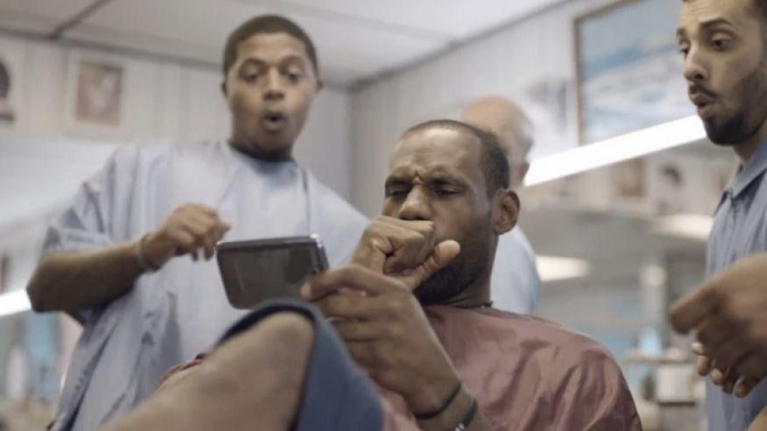 LeBron James staring intently at his phone in a barbershop chair while everyone around him reacts