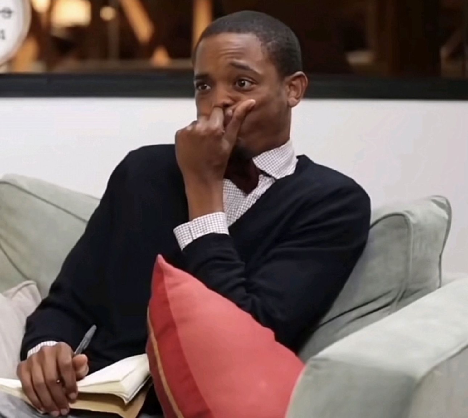 Guy in a bow tie on a couch, hand over his mouth, glancing away mid-thought