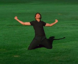 Man on his knees in the middle of a green field, fists up, screaming in triumph