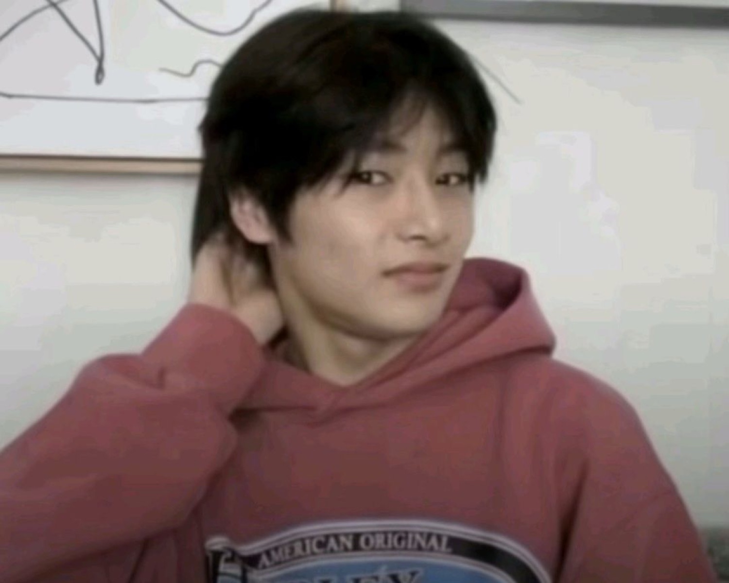 Young man in a red hoodie touching the back of his neck with a sly, coy smile