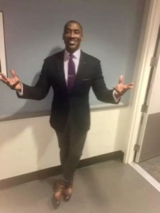 Tall man in a suit spreading his arms wide with a big grin, as if saying 'ta-da'