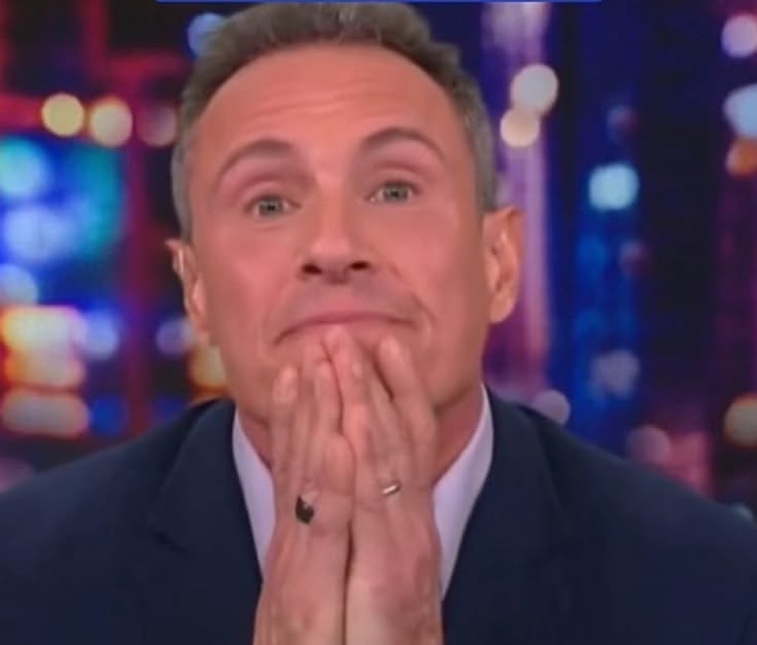 Man on a TV news set pressing his hands to his chin with a tense, contemplative expression