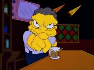 Moe from The Simpsons scowling intensely at a bar with a beer mug, arms crossed