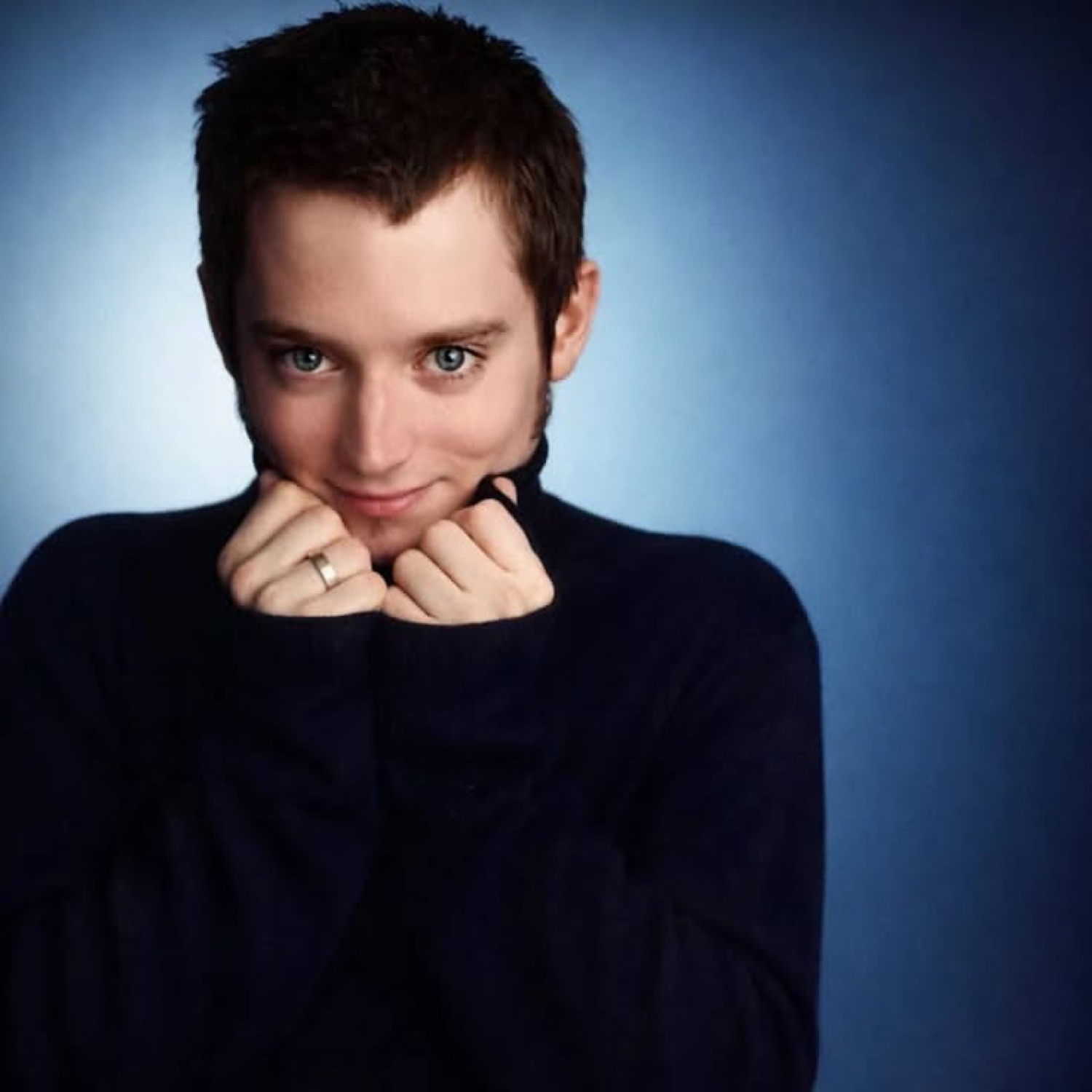 Man in dark turtleneck clasping hands under chin with a sly, pleased-with-himself grin