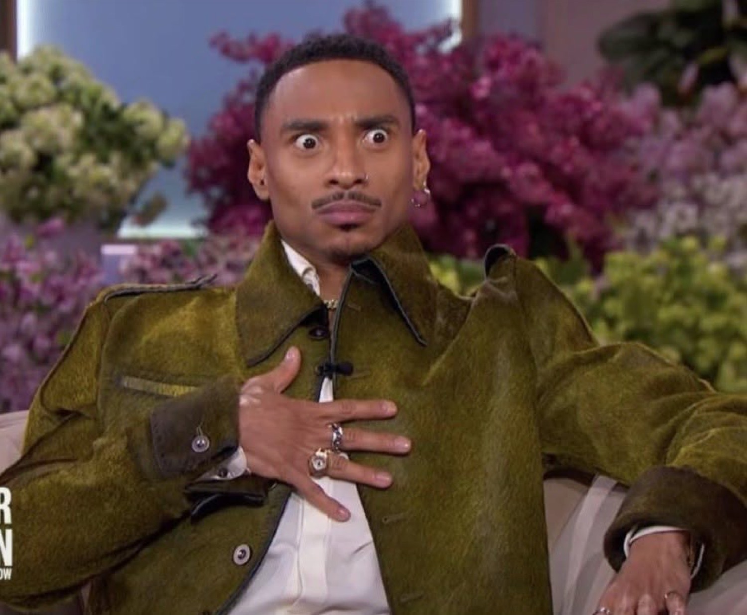 Man on a talk show set with wide eyes and hand on chest in pure disbelief