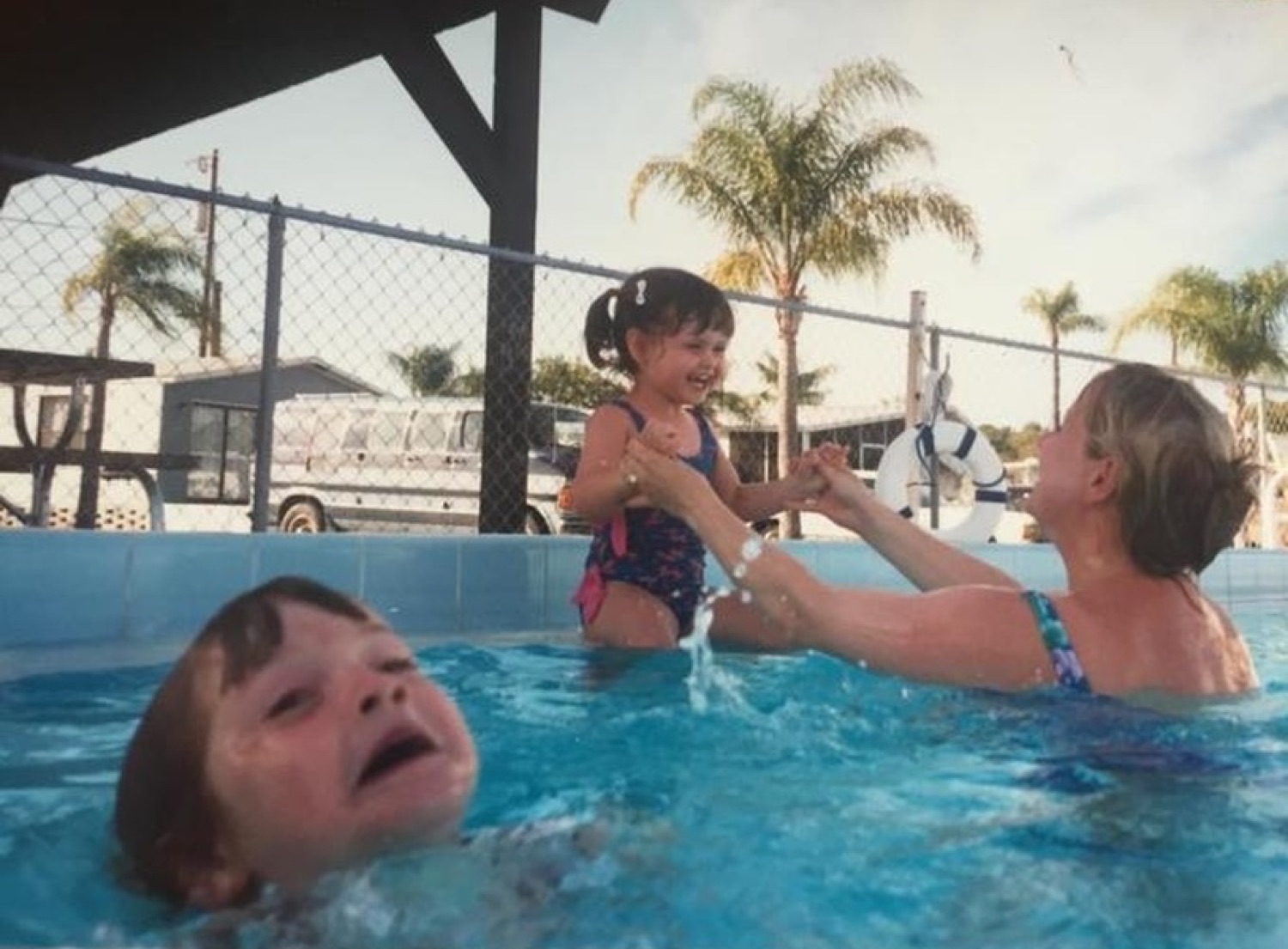 A kid's terrified crying face bobs in the pool while a happy toddler plays in the background