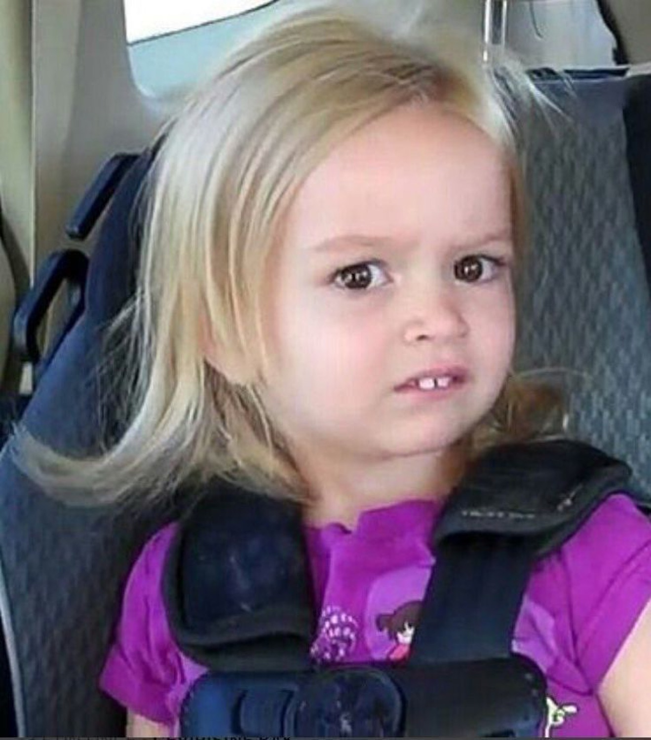 Little blonde girl in a car seat frowning slightly with a skeptical, unimpressed look