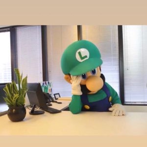 Luigi mascot costume slumped over a desk resting his head on his hand, looking utterly defeated