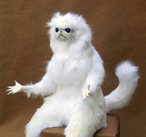 The bizarre white furry cat-monkey figure sitting with an open palm and blank stare