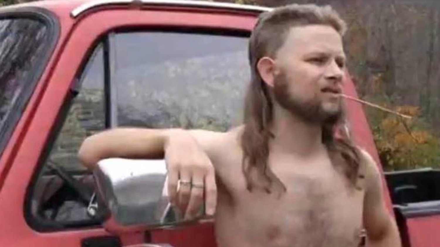 Shirtless man with a mullet leaning on a red truck, chewing a twig with total confidence
