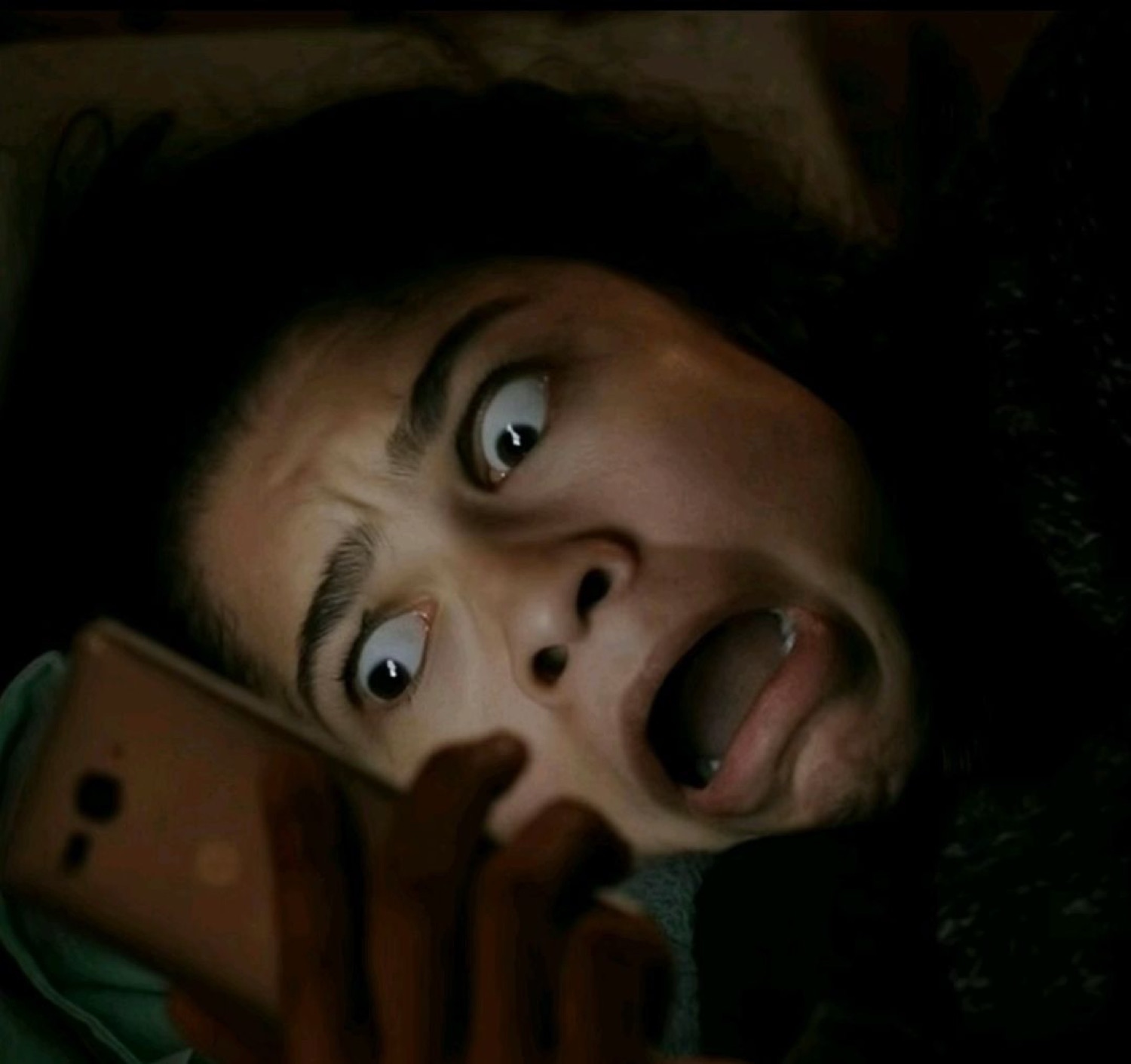 Person lying in bed staring at their phone in the dark with a look of pure horror