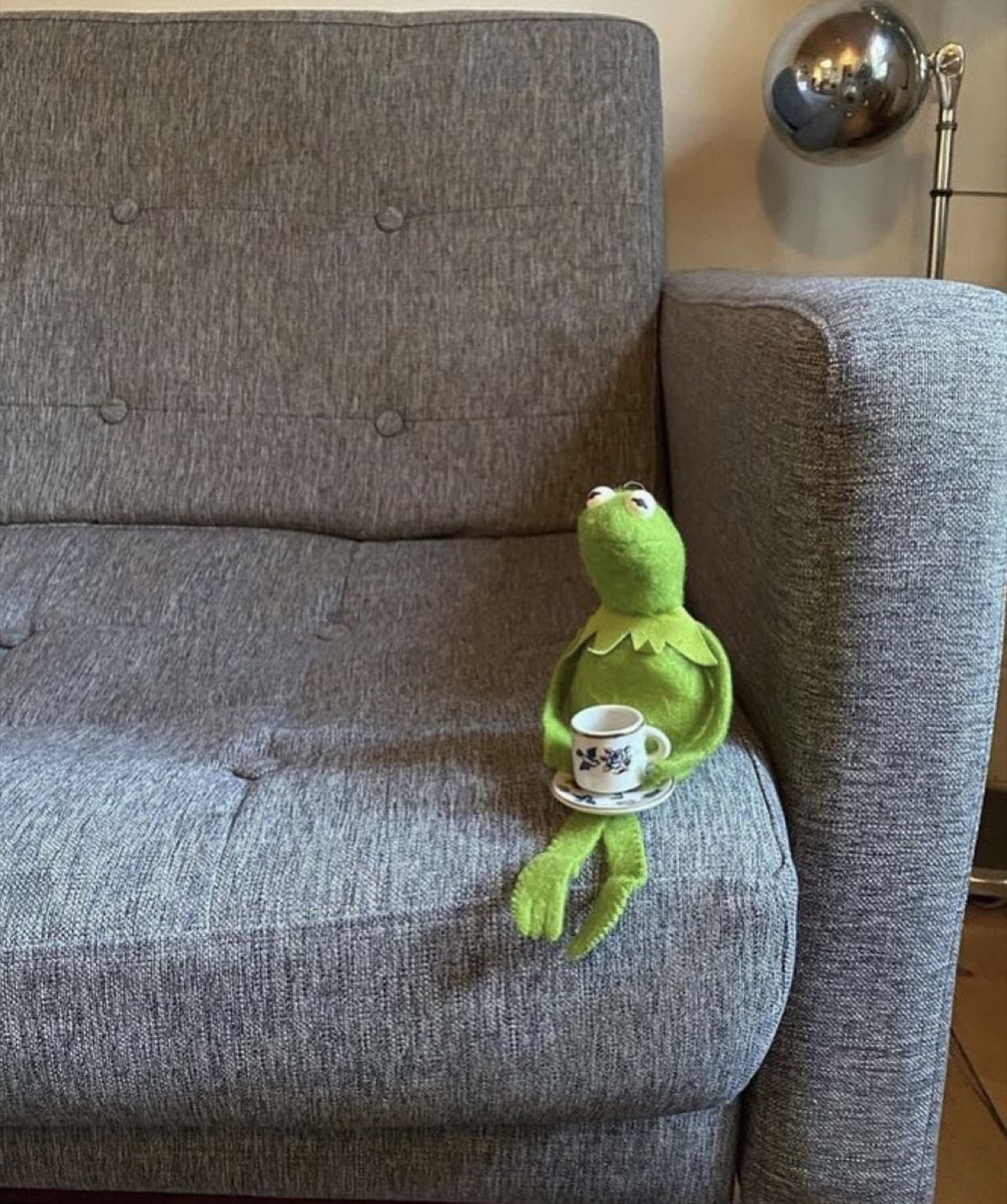 Kermit the Frog sitting alone on a sofa holding a tiny teacup, deeply at peace with it