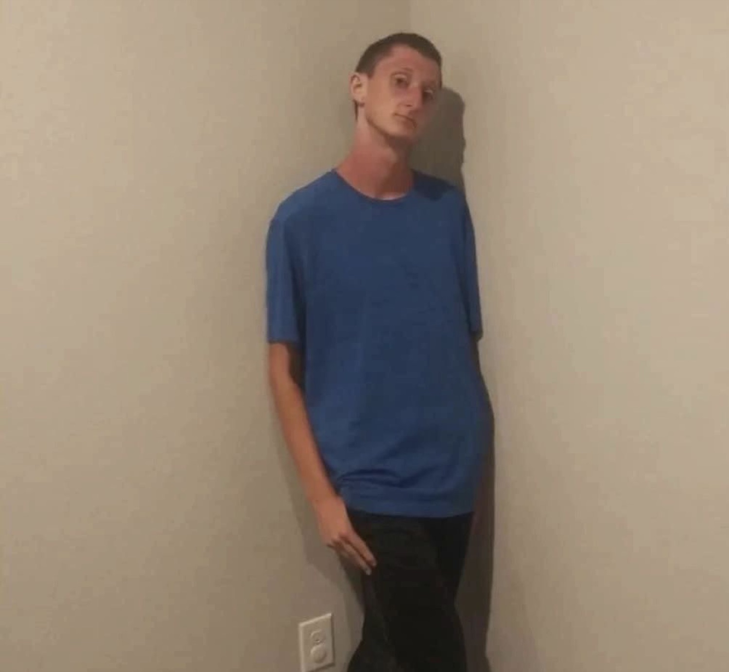 Thin man in a blue shirt leaning into a wall corner with a flat, awkward stare