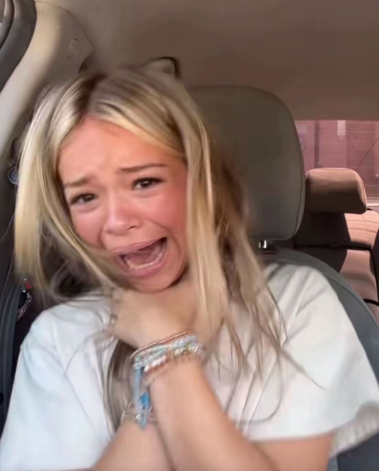 Blonde girl crying in a car with clasped hands, overwhelmed