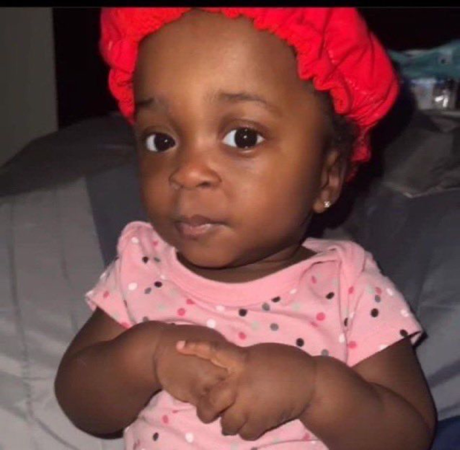 Baby in a red bonnet with hands clasped and a calm, deadpan look