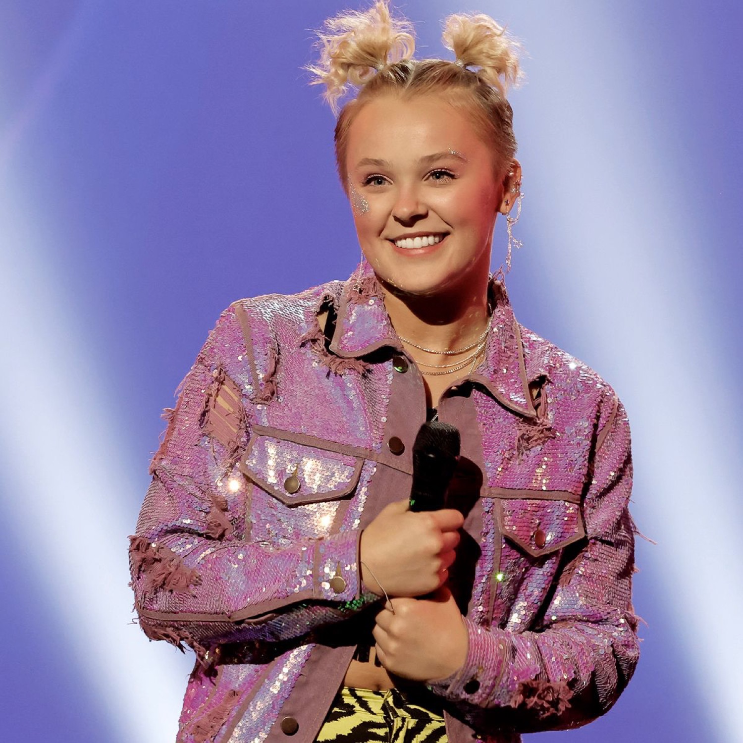 JoJo Siwa beaming on stage in a sparkly pink jacket clutching a microphone