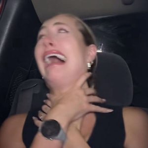 Girl in a car head thrown back, wheeze-laughing with a hand at her throat