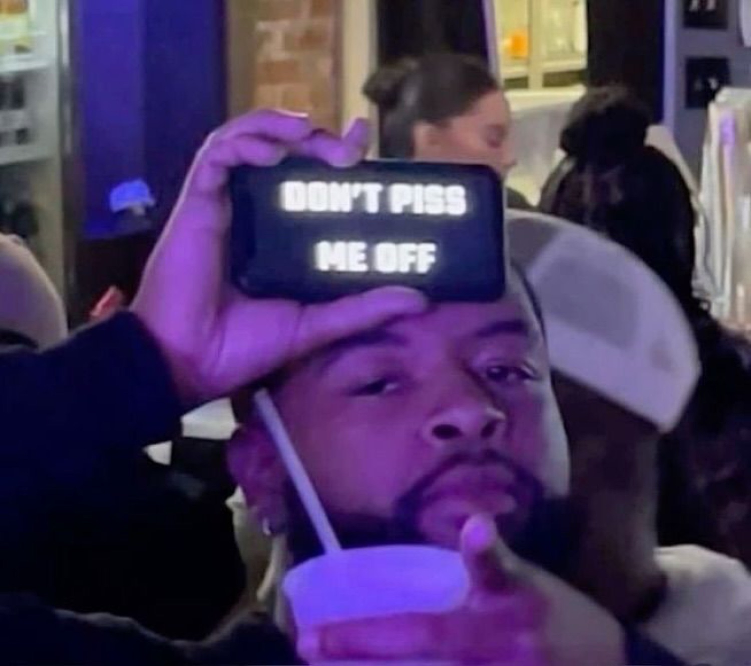 Man holding a phone displaying 'DON'T PISS ME OFF' over his face at a purple-lit party