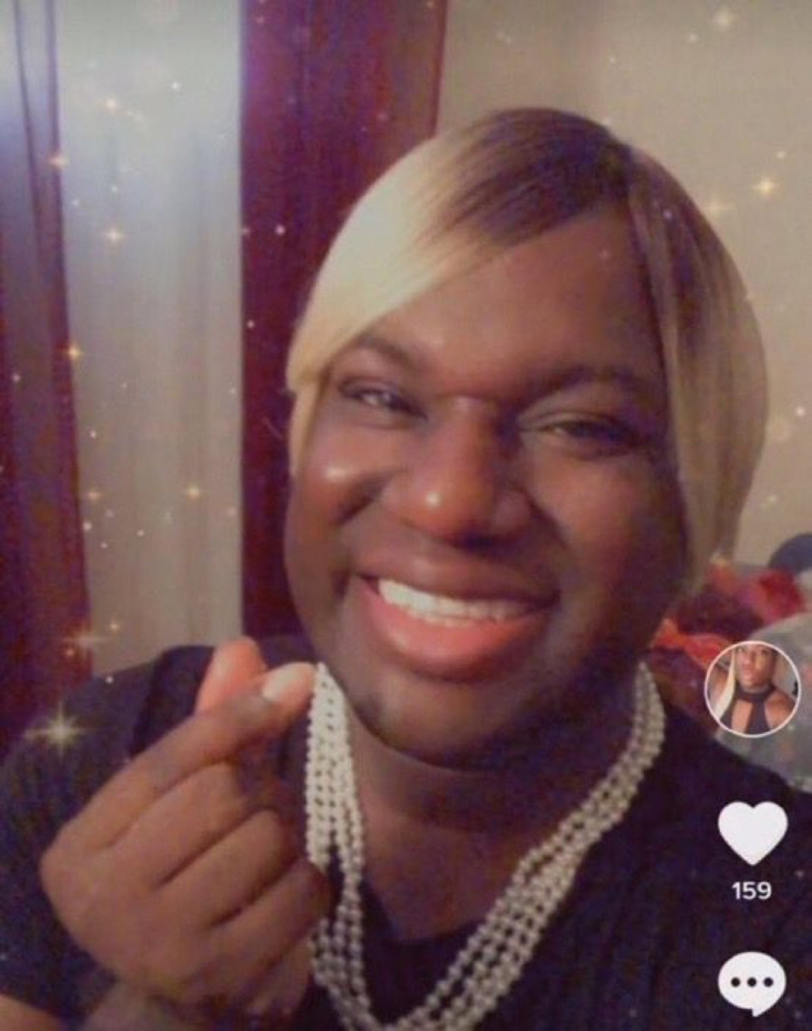 Person in blonde wig and pearl necklace grinning wide with a flirty finger-heart gesture